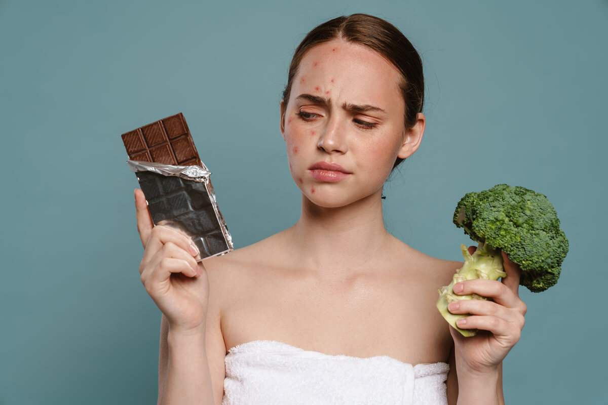 Ginger woman with pimples posing with chocolate and broccoli isolated over blue background