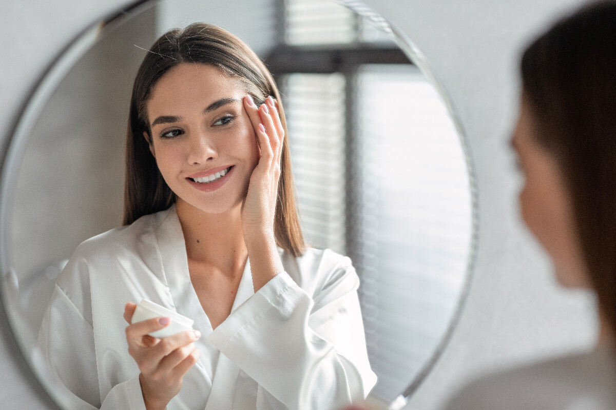 Skincare Concept. Attractive Young Female Looking At Mirror And Applying Moisturising Face Cream, Smiling Millennial Woman Making Beauty Routine At Home, Enjoying Her Skin Condition, Selective Focus