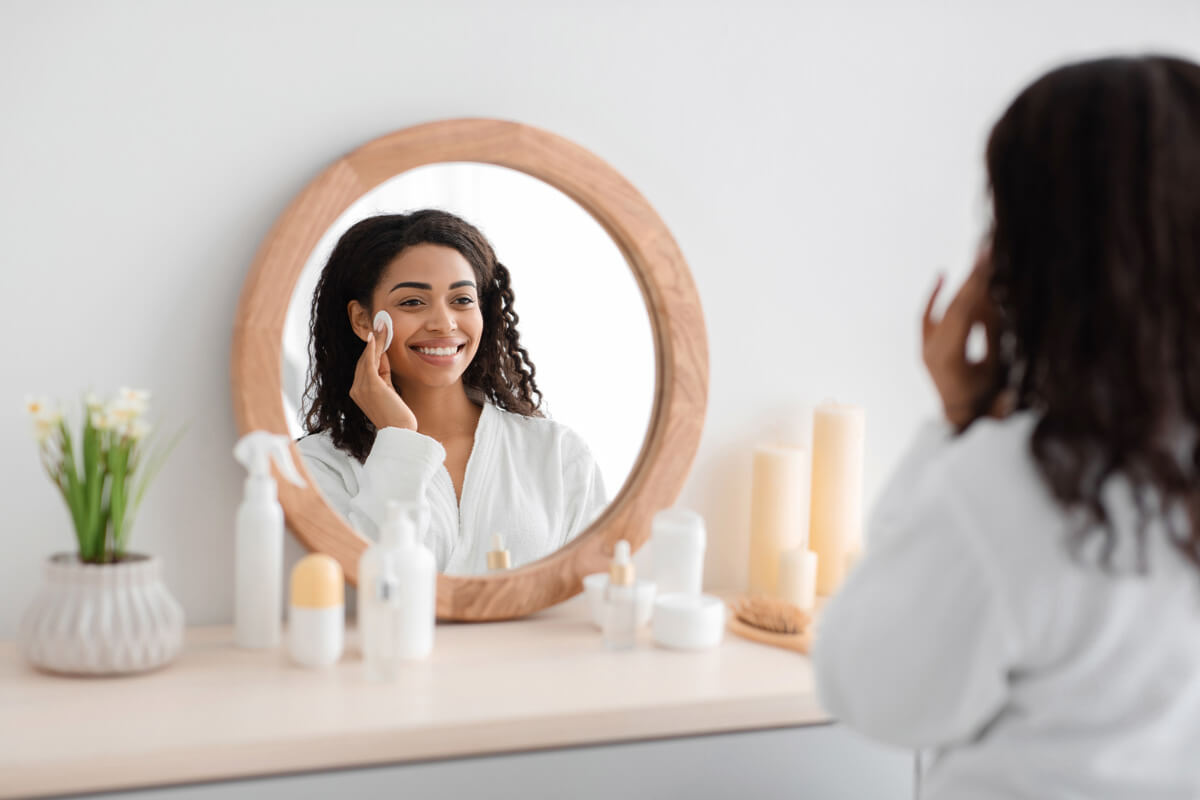 Beautiful skin, makeup remove and routine procedures. Happy millennial african american female in coat applies cream, look in round mirror at table with cosmetics in white bedroom interior