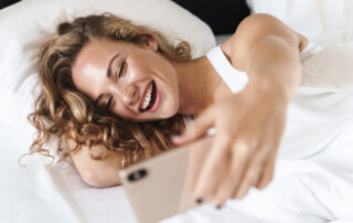 Close up of a happy beautiful young blonde haired woman laying on a pillow in bed, taking a selfie