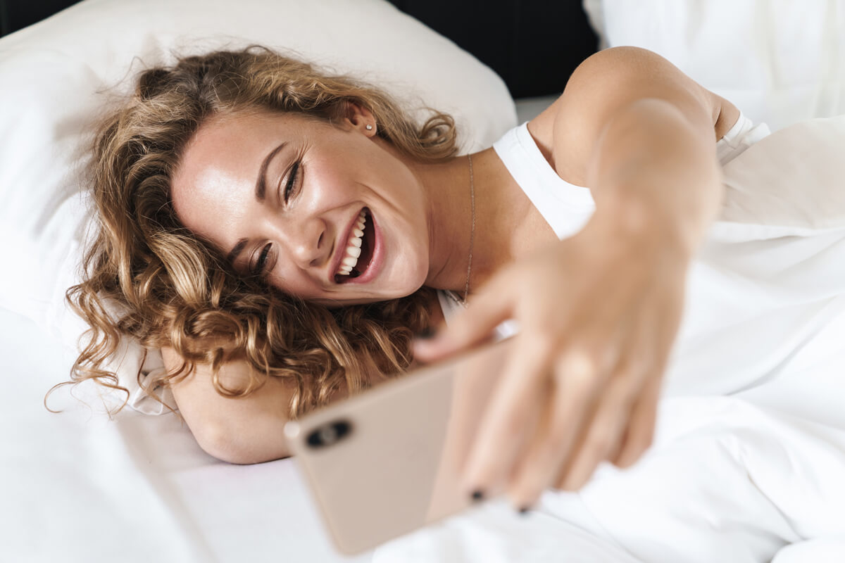 Close up of a happy beautiful young blonde haired woman laying on a pillow in bed, taking a selfie