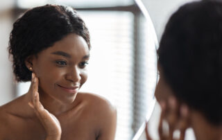 Mirror reflection of cheerful pretty young black woman applying anti-aging or hydrating product on her face after morning shower and smiling, face care cosmetics concept, closeup shot