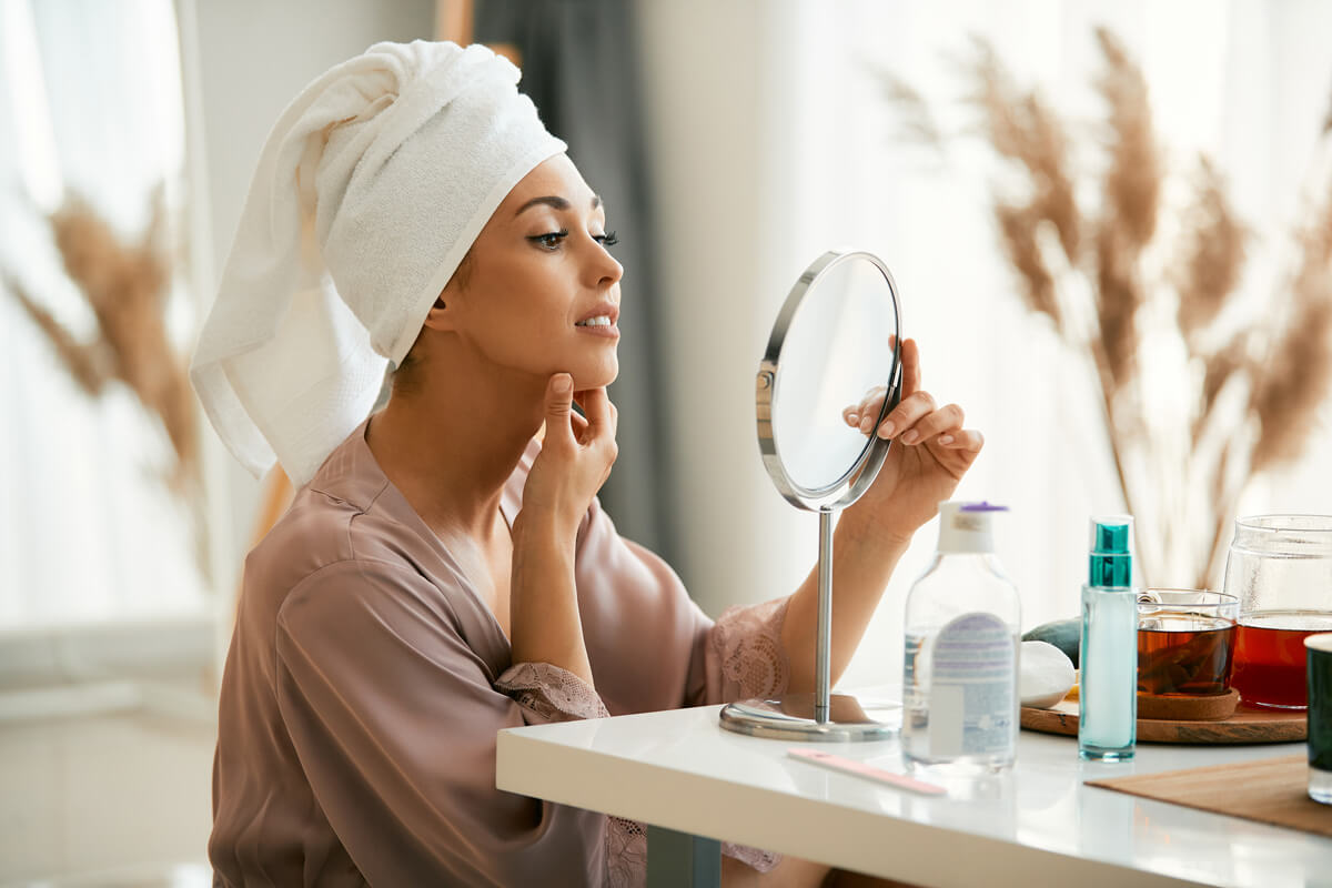 beautiful woman with hair wrapped in a towel looking herself in a mirror at home.