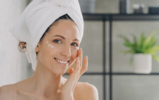 Smiling latina female with naked shoulders moisturizes under eye skin with white face cream, woman in towel apply moisturizing serum, looking at camera in bathroom, enjoying skincare routine at home.