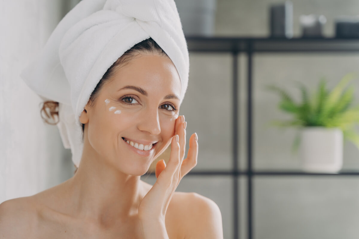 Smiling latina female with naked shoulders moisturizes under eye skin with white face cream, woman in towel apply moisturizing serum, looking at camera in bathroom, enjoying skincare routine at home.